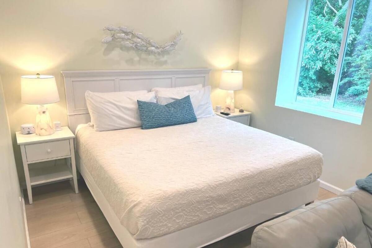 A bedroom with a large bed, two bedside tables with lamps, a decorative wall piece, and a window with a view of greenery.