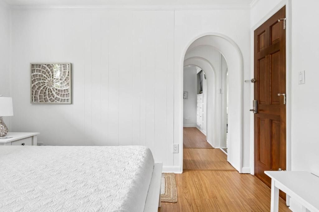 A bedroom with a bed, a wooden floor, a white door, and a hallway leading to another room.