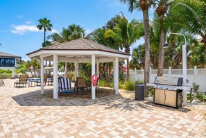A covered outdoor seating area with a grill and poolside chairs. - Siesta Sands 202 | Wake Up Steps from the Gulf (Siesta Key)