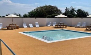 Une piscine entourée d’un petit muret et d’arbres, avec des chaises longues et des parasols.