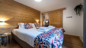 Un dormitorio con techo de madera, una cama con ropa blanca, almohadas de colores, una mesita de noche con vaso de jugo de naranja y una planta colgante.