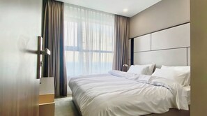 Moderni hotellihuone, jossa on suuri sänky, yöpöytä, ikkuna, jossa on läpikuultavat verhot, ja neutraali värimaailma.