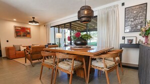 A dining area with a wooden table and chairs, a large window with curtains, and a hanging light fixture. - Modern 3BR Pool & Gym in Nueva Escondida (San Miguel de Allende)