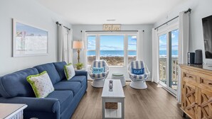 Un salon avec un canapé bleu, deux fauteuils ornés de coussins aux motifs marins, une table basse et une vue sur l’océan grâce à de grandes fenêtres.