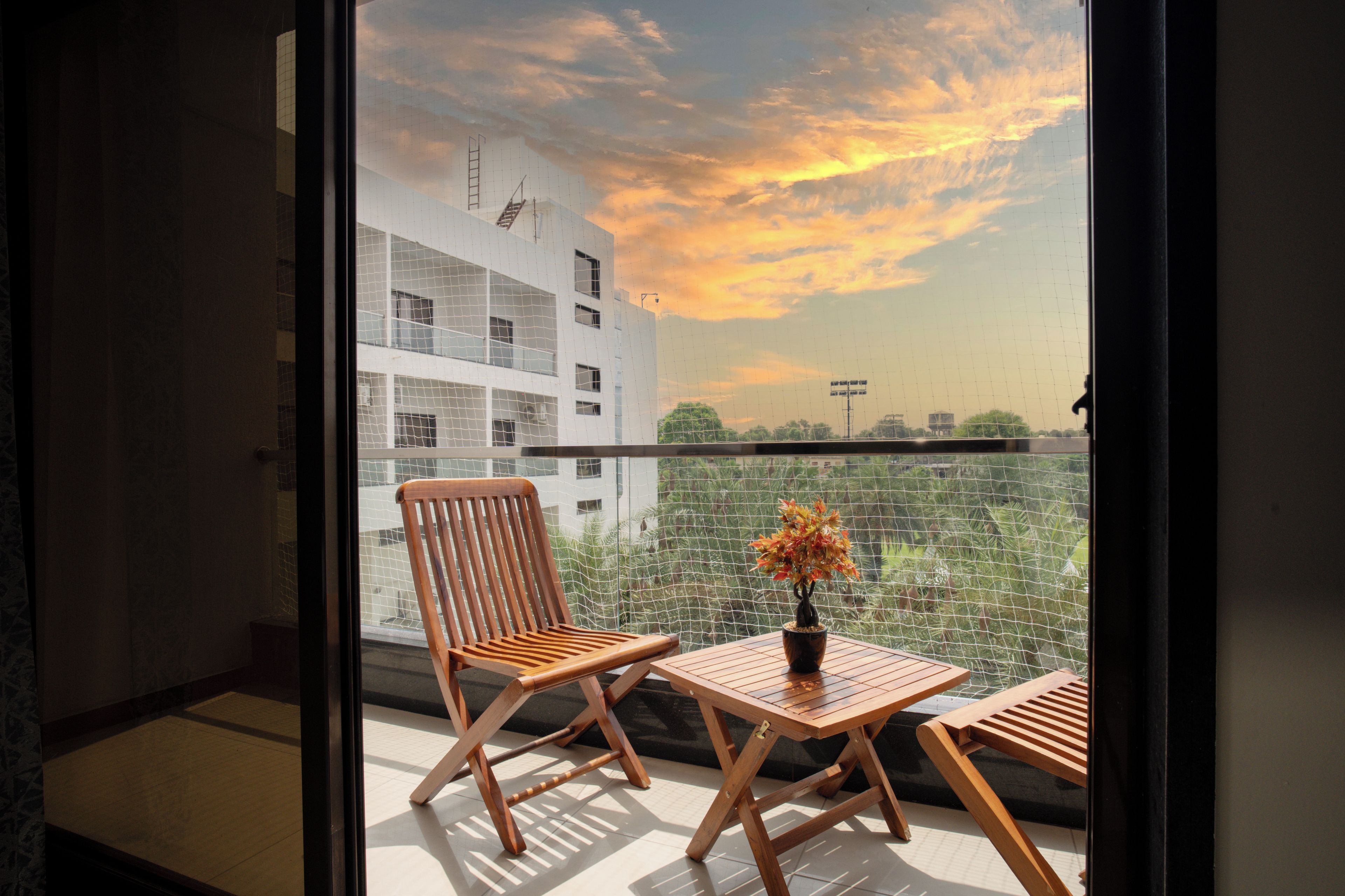 A balcony with a wooden chair and table, a potted plant, and a view of a building and sky.