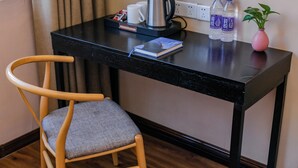 Une chaise en bois avec un coussin gris, un bureau noir avec une bouilloire, deux bouteilles d’eau, un vase rose avec une plante et un livre.