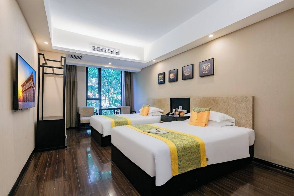 A hotel room with two beds, a desk, a chair, a window with a view of trees, and framed pictures on the wall.