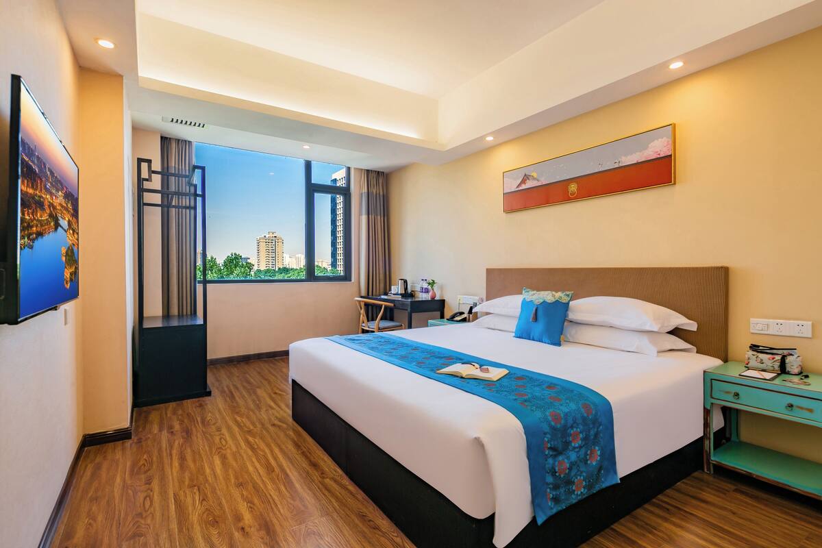 A hotel room with a large bed, a desk, a chair, a nightstand, and a window with a view of buildings.
