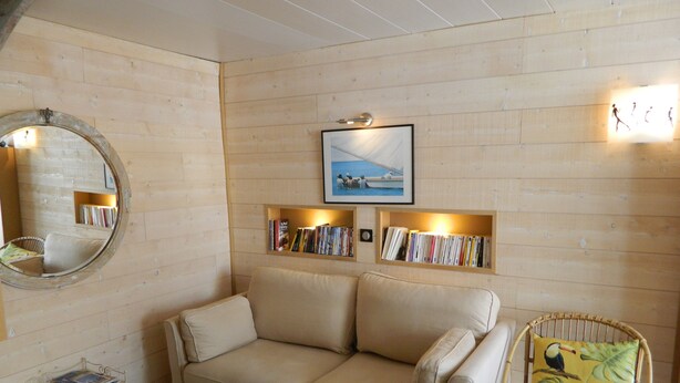A room with a beige sofa, a wicker chair with a tropical print cushion, a round mirror, and two wall-mounted bookshelves with books and framed pictures. - Nice little house in a good location to forget your car during the vacations. (Paris)