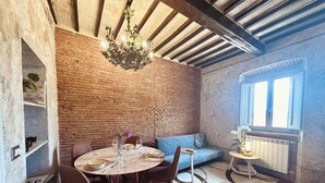 Uma sala de jantar com parede de tijolos, um candelabro, uma mesa redonda com cadeiras, um sofá azul e uma janela com persianas.
