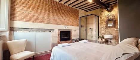 A bedroom with a brick wall, a fireplace, a bed, a chair, and a shower area.