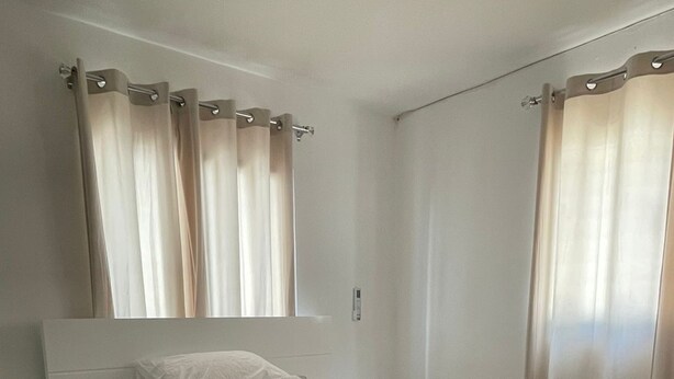 En snyggt bäddad säng med ett vitt täcke och kuddar, ett sängbord med en lampa och ett fönster med gardiner.