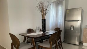 En matplats med ett bord, stolar, en vas med torkade växter, ett kylskåp och ett fönster med gardiner.