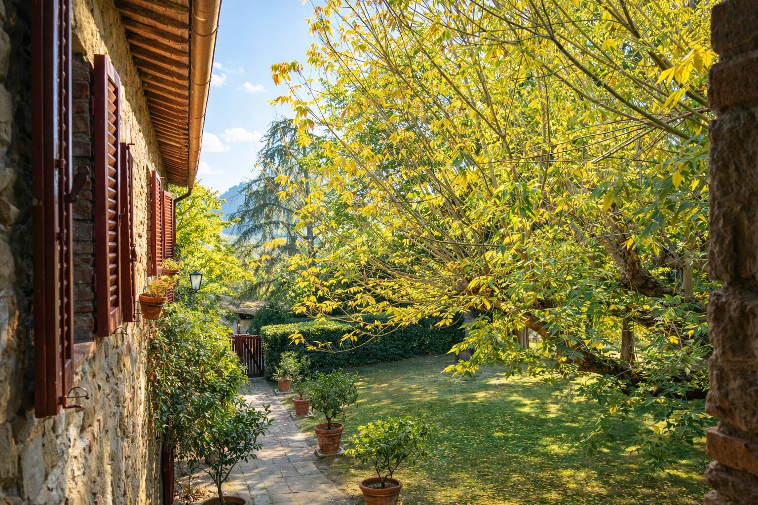 Un bâtiment en pierre avec des volets rouges, un chemin pavé et un jardin agrémenté d’arbres et de plantes en pot.