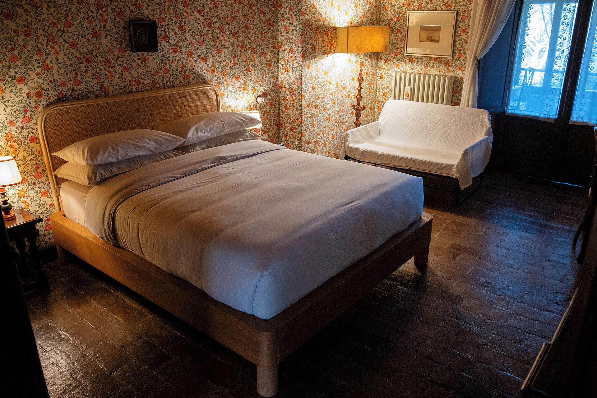 Una cama doble con cabecera, una mesita de noche con lámpara, una silla y una ventana con cortinas.