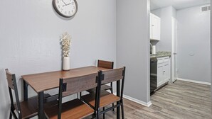 Una zona de comedor con una mesa y sillas de madera, un reloj de pared y un jarrón con flores.