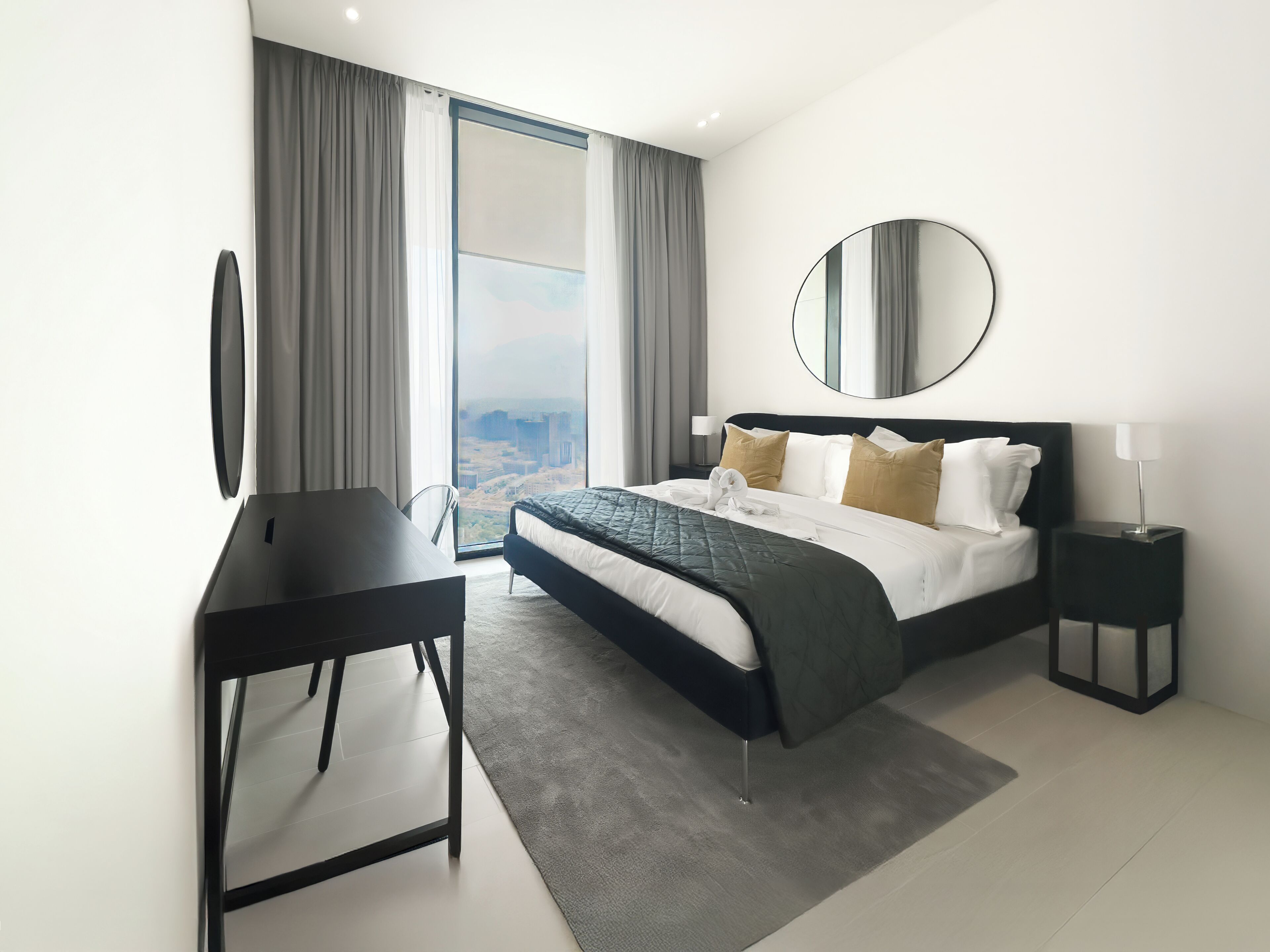 A modern hotel room with a large bed, a desk, a round mirror, and a view of a cityscape.