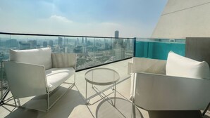 Un balcón con sillas blancas y una mesa redonda, con vista a un paisaje urbano de edificios modernos.
