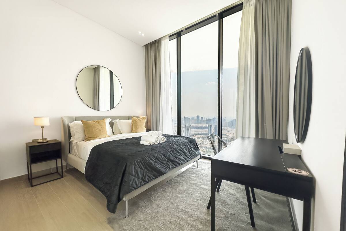 Een moderne hotelkamer met een groot bed, een bureau, een ronde spiegel en uitzicht op de stad door grote ramen.