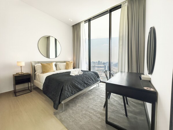 Ett modernt hotellrum med en stor säng, ett skrivbord, en rund spegel och utsikt över stadsbilden genom stora fönster.