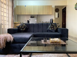 Ein Wohnzimmer mit einem dunkelblauen Sofa, zwei gemusterten Kissen, einem Glastisch und einer Küche im Hintergrund.
