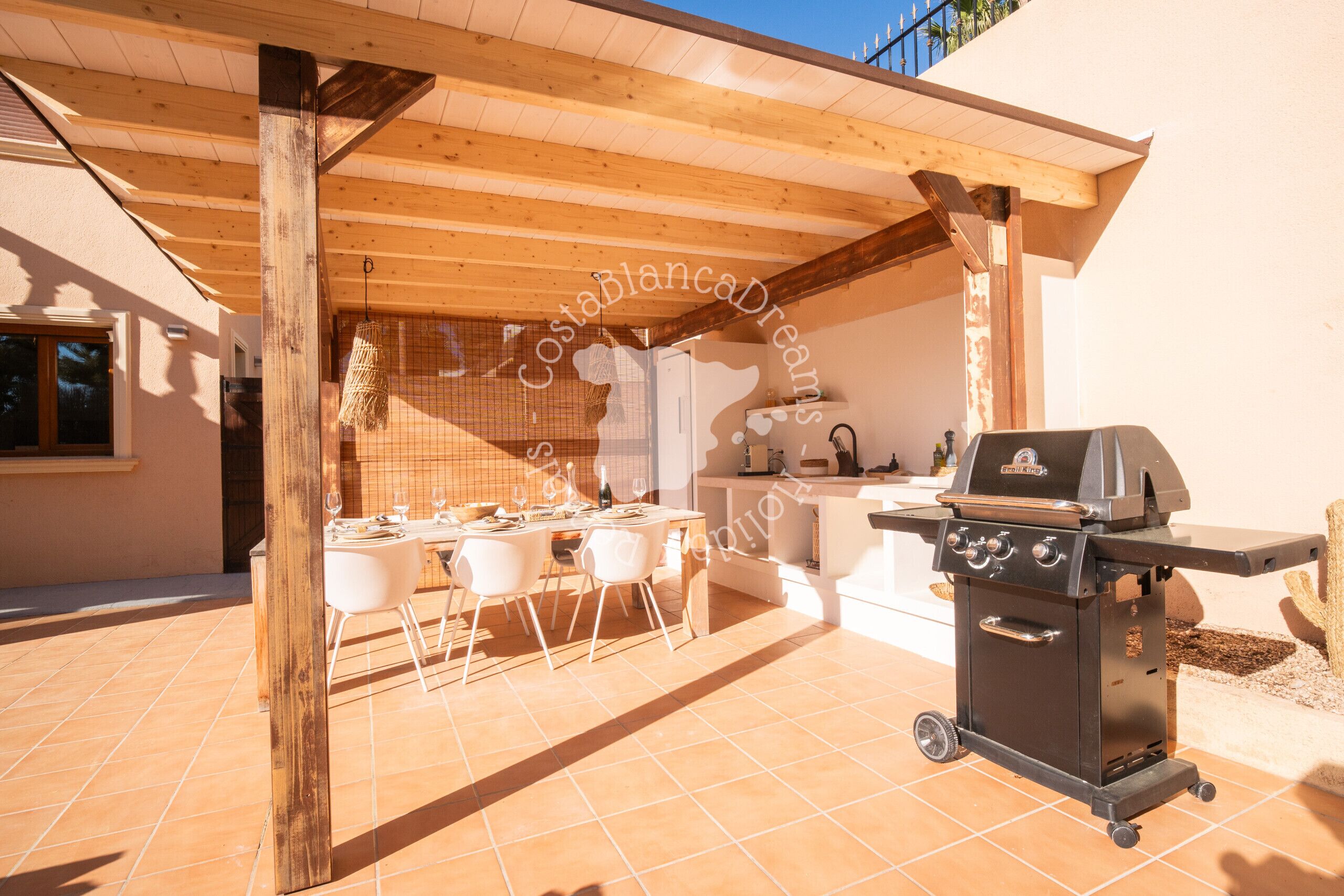 Une terrasse extérieure dotée d’une pergola en bois, d’un barbecue et d’un espace repas avec des chaises et une table.
