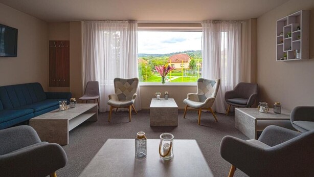A living room with a sofa, armchairs, a coffee table, and a view of a house through a window. - Hotel Aréna (Brumov-Bylnice)