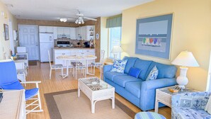 Sala de estar con un sofá azul, mobiliario de mimbre blanco, una cocina al fondo y un ventilador de techo.