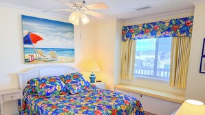 Un dormitorio con cama, ventilador de techo, ventana con cortinas, mesita de noche y un cuadro de una playa.
