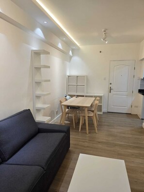 Ein modernes Wohnzimmer mit einem dunklen Sofa, einem weißen Couchtisch, einem Essbereich mit Holztisch und Stühlen sowie einer weißen Tür.
