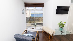 A room with a view of the sea, a wooden bench, a chair, a small table with a plant, and a TV mounted on the wall.