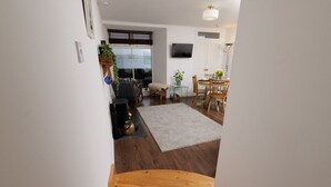 A modern living room with a wooden staircase, a dining area with a round table, and a large window with blinds.