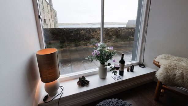 A room with a large window, a lamp, a vase with flowers, a bottle, and a small house model on the windowsill.