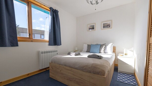 A bedroom with a wooden bed, a radiator, a window with curtains, a bedside table, and two framed pictures on the wall.