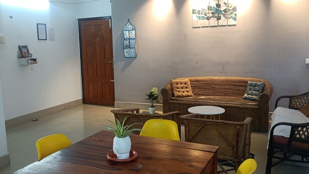 A dining area with a wooden table, yellow chairs, a wicker sofa, and a small round table. - KK HOMES GUWAHATI (Guwahati)