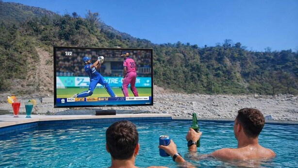 A poolside with a television displaying a cricket match, a bottle, and two people in the water. - Natures Crown Resort (Dehradun)