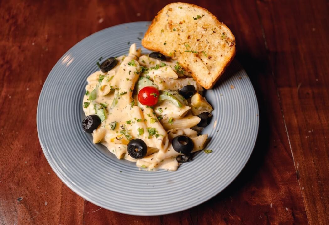 Un plato de pasta con aceitunas negras, un tomate cherry y una rebanada de pan sobre un plato azul.