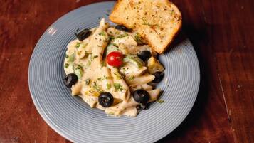 Un plato de pasta con aceitunas negras, un tomate cherry y una rebanada de pan sobre un plato azul.