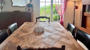 A dining room with a wooden table, chairs, a hanging light fixture, a mirror, and a window with curtains. - Family vacation home facing the beach on the edge of the GR34 (Porspoder)