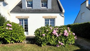 A two-story house with a white exterior, grey roof, and a small front garden with pink flowers. - Family vacation home facing the beach on the edge of the GR34 (Porspoder)