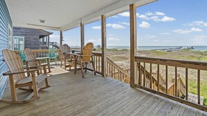 Een houten terras met schommelstoelen en een tafel, met uitzicht op een strand en een pier.