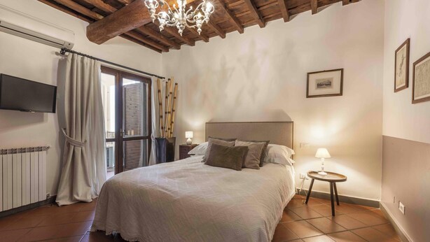 Makuuhuoneessa on sänky, televisio, kattokruunu, ikkuna, jossa on verhot, sekä pieni yöpöytä, jossa on lamppu.