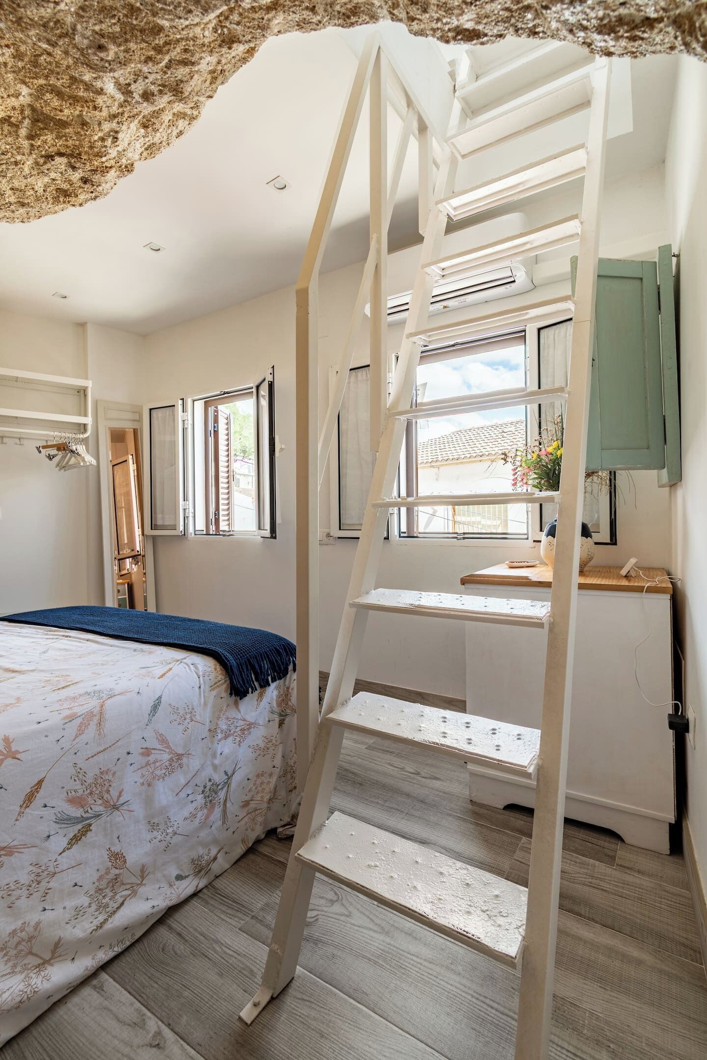 A bedroom with a bed, a desk, a ladder, and a window with a view.