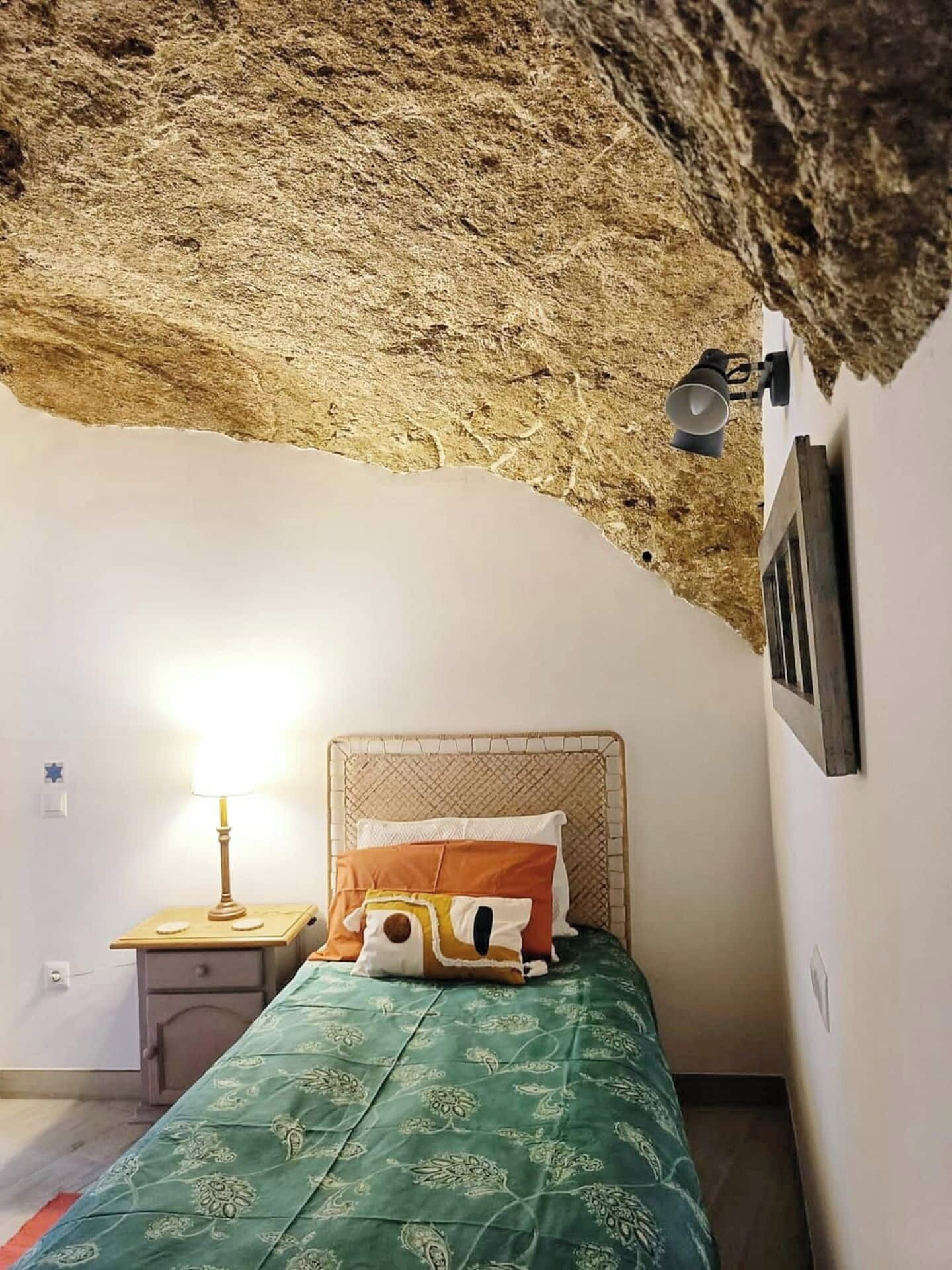 Een slaapkamer met een plafond dat doet denken aan een grot, een bed met beddengoed met een patroon, een bedlampje en een klein nachtkastje.