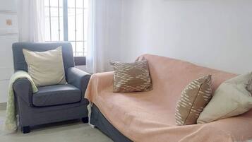 A living room with a grey armchair, a sofa with a peach-colored throw, and a window with white curtains.