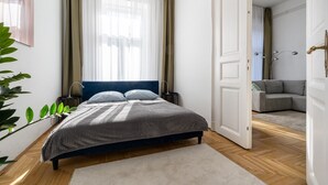 A bedroom with a bed, a bedside table, a lamp, a plant, and a view into another room. - AVATAR Suite (Budapest)