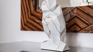 A white abstract sculpture on a reflective surface with a wooden geometric wall and a plant in the background. - AVATAR Suite (Budapest)