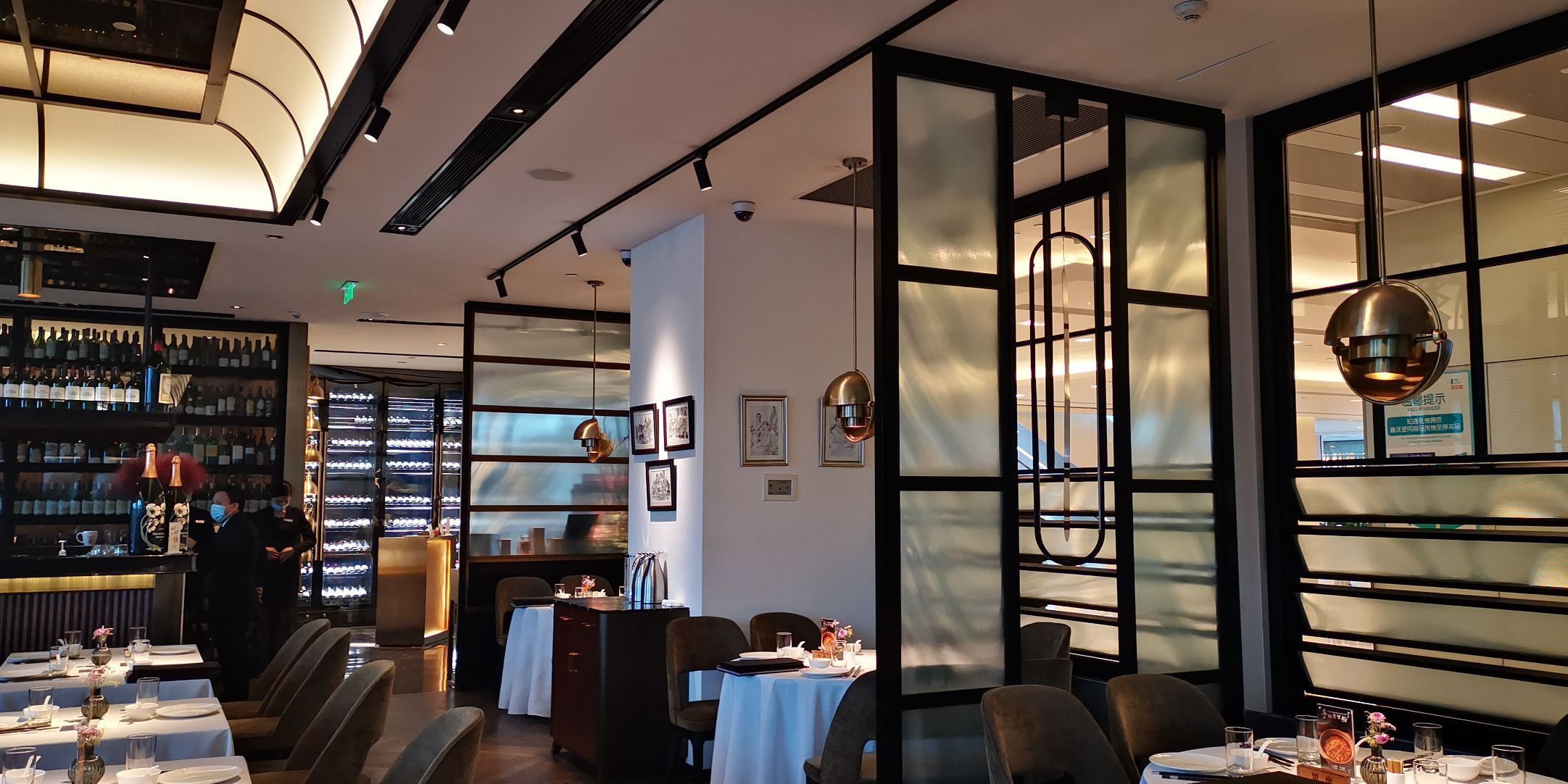 A restaurant interior with tables set for dining, a bar area with bottles, and a view through a glass partition.
