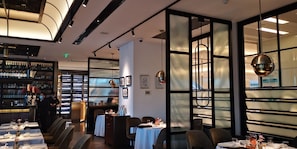 Interior de restaurante con mesas dispuestas para comer, una zona de bar con botellas y vistas a través de un panel de vidrio.