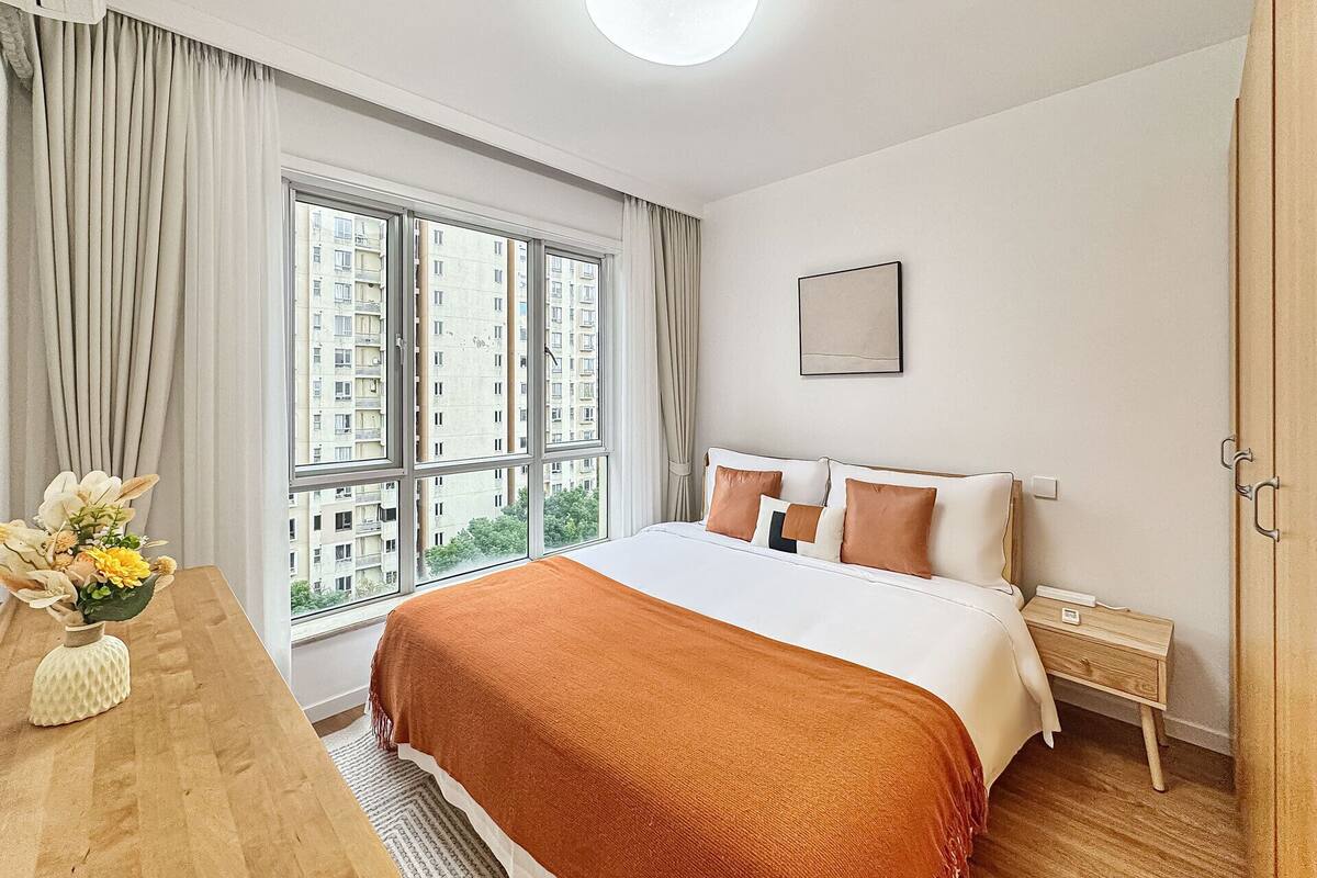 Una habitación de hotel con una cama grande, una mesita de noche, cortinas en la ventana e iluminación en el techo.